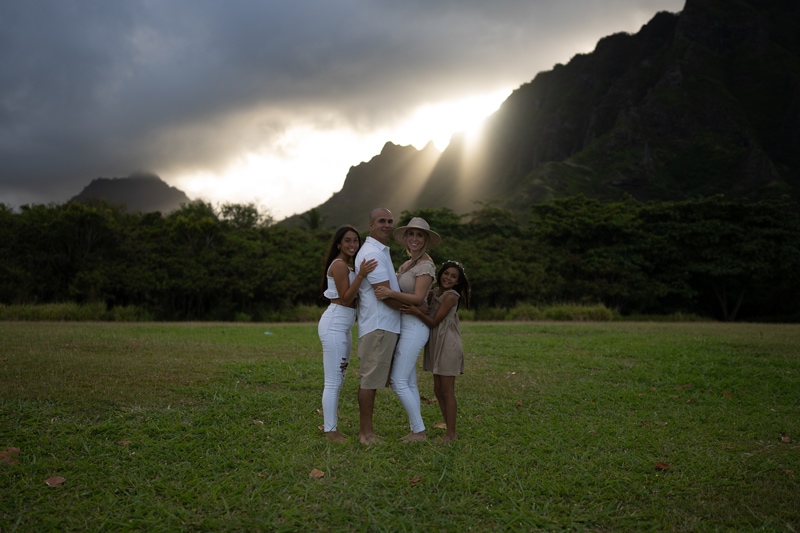 dark unedited image, a family of four embrace in lush green grass near tropical mountains, the light shines on them