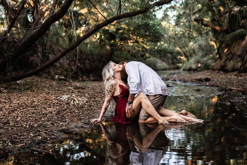 Photographer education, woman sits in quiet stream in forest in dress, man leans into her knees in the water