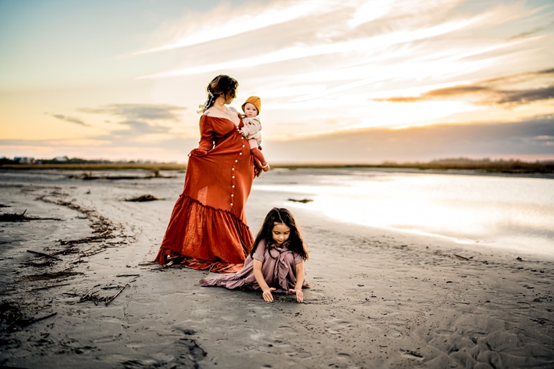 Mother holds baby in a dress, while daughter plays in the sand beside her, they are at the beach