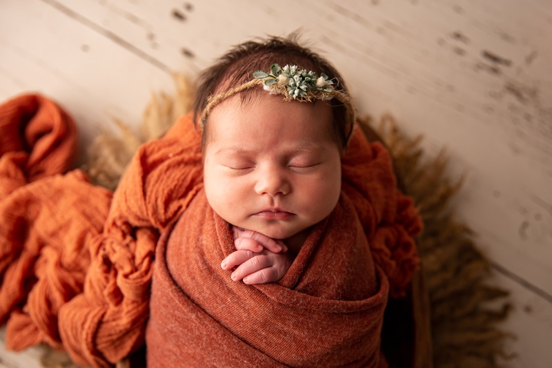 Dark image, a baby lays sleeping swaddled in woven blankets, a flower headband in her hair