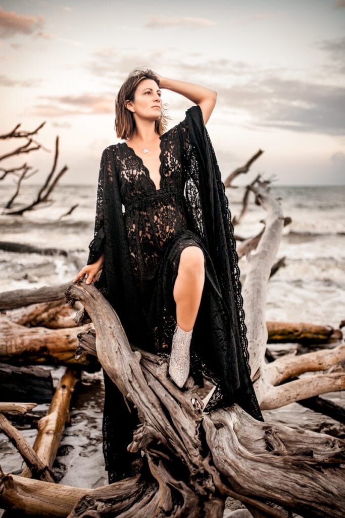 Victoria Armstrong stands on large drift wood near the ocean in a black laced beach cover uo