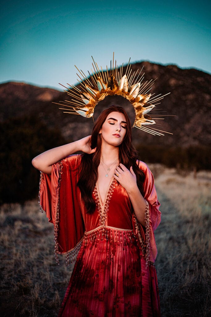 Woman stands in field before mountains, wearing gown and golden crown